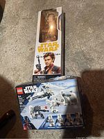 Photo of two sealed items: a Star Wars Lego set with stormtrooper characters and a Han Solo action figure with writing on the box.