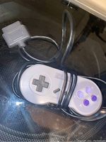 One Super Nintendo Entertainment System controller with attached cable and connector, shown on a reflective surface.