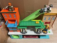 Photo showing the full TMNT play set including orange multi-level building, green-tiled roof structure, blue slide, beige building, and three TMNT figures placed around the set.