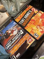 Photo showing two PlayStation 2 game cases: Tony Hawk's Pro Skater 4 and Tony Hawk's Underground 2 on a surface near a steering wheel accessory.