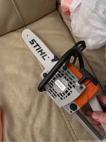 Plastic toy Stihl chain saw with orange, gray, and white coloring, featuring Stihl branding on blade and body, shown on beige couch