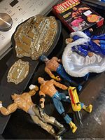 Photo of wrestling lot including UFC belts, three action figures, blue and white robe, and WWE VHS tape on dark surface.