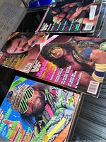 Four wrestling magazines laid out showing cover images of wrestlers including one with face paint and championship belt, one with sunglasses, one biting finger, and one with serious expression