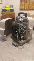 Full view of Bell and Howell vintage film projector with dust cover placed beside it on a wooden surface.