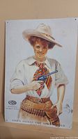 Front of the tin sign showing the detailed vintage illustration of a woman holding a Colt revolver.