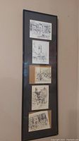 Full view of the tall black frame with five black and white ink sketches arranged vertically, showing architectural street scenes.