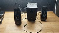 Two black Logitech satellite speakers and one subwoofer on wooden surface, wired with green 3.5mm audio jack cable. Clear Logitech logos and speaker drivers visible.