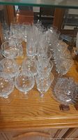 Fourteen clear crystal glassware pieces including various stemmed glasses and a round covered dish with a pinwheel cut pattern arranged on a wooden surface.