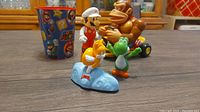 Photo showing 5 Super Mario themed items: Mario figurine in white outfit, Donkey Kong on kart, green Yoshi, yellow Paratroopa on kart, and a Super Mario plastic cup with character designs.