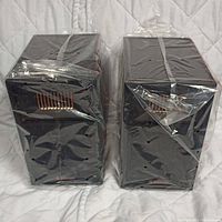 Two Donner bookshelf speakers, black, wrapped in plastic, front and sides partially visible with copper coil elements showing on the rear side of each speaker.