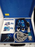 Open carrying case displaying shockwave therapy device components: handheld applicator with attached cables, four interchangeable treatment heads, and user manual.