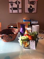 Overall lot showing wicker basket, metal basket with faux fruit, purple vase, white cylindrical vases, and part of furniture in background.
