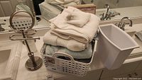 Photo of white and beige bathroom towels stacked in a plastic basket with round tabletop makeup mirror and white plastic waste bin nearby