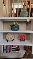 Shelf showing green ceramic vase on the top, blue woven basket with faux plant in the middle, two decorative plates on stands below, and several books on the bottom shelf.
