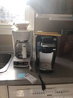 Two coffee makers side by side on countertop: a white Mr. Coffee drip coffee maker with glass carafe and Keurig black single-serve coffee maker with power cord and manual.