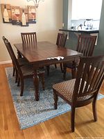 Full view of wood dining table and six matching chairs with patterned upholstery, arranged on blue patterned area rug.