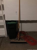 Full view of broom, waste can, orange extension cord, and garden hose in garage setting