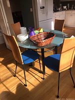 Round kitchen table with frosted glass top surrounded by four wood chairs with blue seat cushions in a kitchen setting, illuminated by natural light.