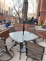 Photo showing square outdoor glass-top table with metal frame surrounded by four matching mesh and metal patio chairs and umbrella stand in the middle.