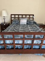 Front view of queen size wooden bed with blue-gray comforter, pillows, and nightstand with lamp nearby.