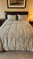 Front view of queen bed made up with beige floral comforter and light blue pillows and sheets against dark wood headboard.