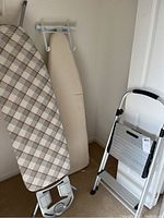 Photo of two ironing boards, one with plaid fabric cover and one with beige fabric cover, and a Cosco step ladder with aluminum steps and black hand grips, leaning against white wall.