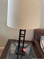 Black metal table lamp with rectangular base, vertical geometric cut-out design and white cylindrical fabric shade, photographed on glass table near a couch.
