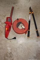Photo of electric hedge trimmer, extension cord on orange cordwheel, and black loppers with orange handles on concrete floor