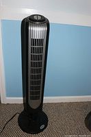 Full front view of black and silver Holmes tower oscillating fan standing upright on carpeted floor against a blue wall.