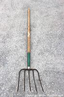 Full length of vintage pitch fork laid flat on concrete surface, showing wooden handle and metal five-pronged fork head