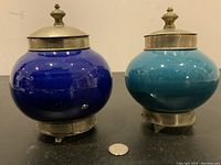 Front view of two pottery jars, one dark blue and one turquoise, with metal fitted bases and lids, displayed on black surface next to a coin for scale.