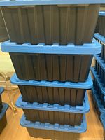 Stacked view of four dark blue heavy-duty plastic storage containers with bright blue lids on a wood floor, showing the containers are stackable.
