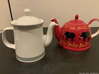 Side view of white Dansk teapot with gray band next to red teapot with black elephants and gold leaf decoration on a white background.