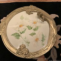 Top-down photo of round brass Art Deco frame containing a signed watercolour of white daisies, with brass figure positioned on frame edge.