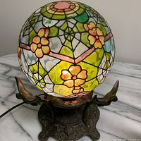 Front angle showing colorful stained-glass floral pattern on glass ball lamp lit and resting on ornate metal base.