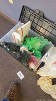 Black plastic storage bin containing approximately 10 Christmas ornaments and decorations including bottle brush trees, wrapped glittery ornaments, green bubble wrap, and white boxes.