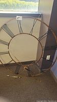 Large round metal skeleton frame wall clock approximately 50 inches in diameter leaning against a wall near a window. Clock has Roman numerals and visible hour and minute hands.
