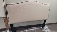 Photo of a beige fabric upholstered headboard with dark metal nailhead trim and black support legs.