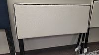 White upholstered full sized headboard with black nailhead trim and black legs against a grey wall.