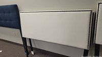 Full sized white upholstered headboard with nailhead trim shown standing against a wall in a room.