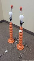 Pair of orange retro table lamps with stacked spherical bases and metal harp, no shades, placed on carpeted floor.