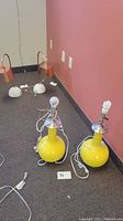 Two yellow modern mid century style table lamps on floor, one with standard bulb one with CFL bulb, cords visible