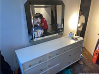 Front and angled view of white dresser with 8 drawers topped with various items, including a roll of toilet paper and the matching mirror mounted above.