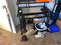 Black glass and metal desk with pull-out keyboard shelf and lower shelf, showing some surrounding clutter on carpeted floor.