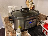 Front and side view of InstaPot digital slow cooker on countertop revealing control panel and glass lid with handle.