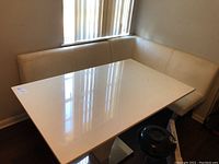 White Formica kitchen table with L-shaped cream vinyl corner seating placed beside it.