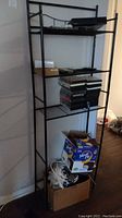 Side angle view of black metal shelving unit with DVDs, electronics, boxes of cables underneath