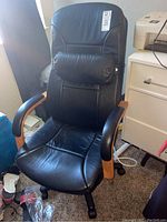 Photo of black vinyl office chair with wooden armrests, padded headrest pillow, and five caster wheels, placed indoors by a window next to a white drawer unit.
