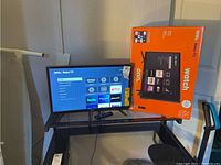 Onn Roku TV powered on and displaying streaming interface, placed on a black stand with remote in front and orange box next to it.