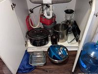 Kitchen cabinet with various kitchen items including Crux crockpot, metal bowls, glass baking dish, blender, and utensils.